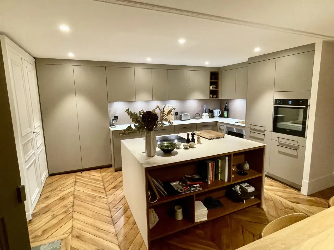 Une cuisine ouverte moderne avec un &icirc;lot central, des placards gris, un parquet en chevrons et une d&eacute;coration florale.