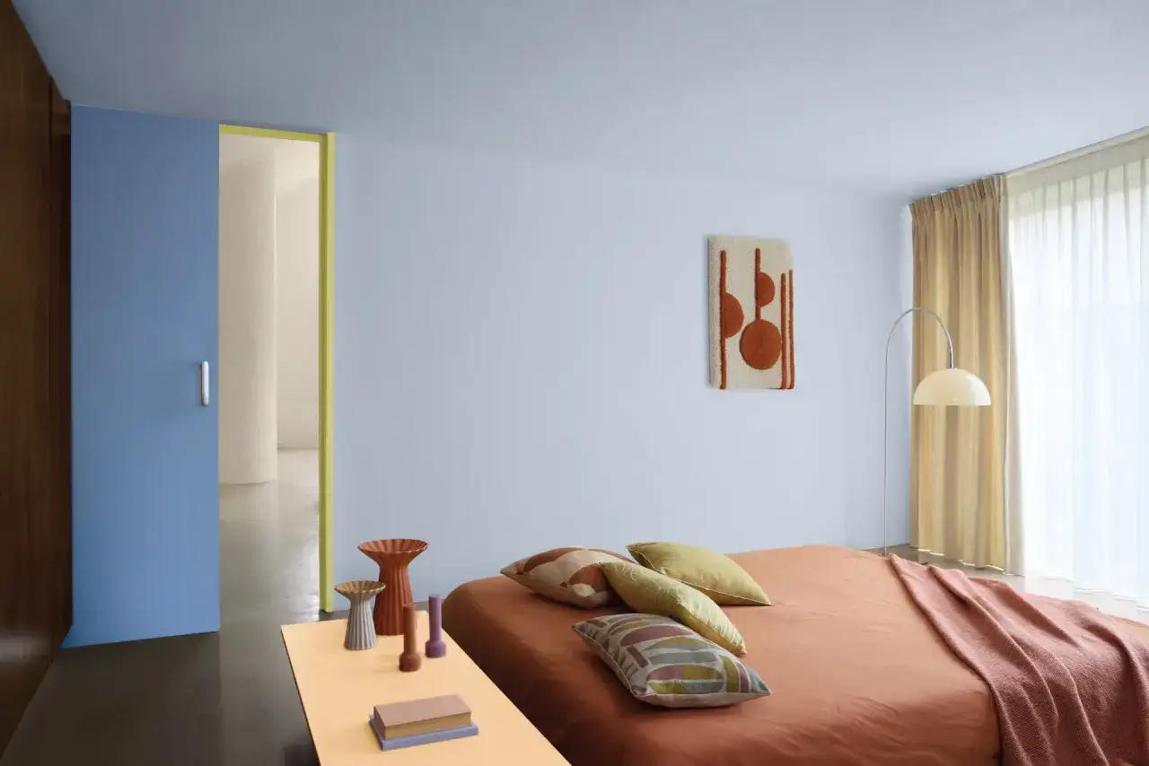 Une chambre aux murs bleu p&acirc;le, pr&ecirc;te &agrave; peindre. Un lit orange avec des coussins color&eacute;s, une porte bleue et des objets d&eacute;coratifs sur une table.
