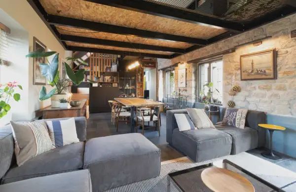 Salon cosy d'une maison de pêcheur rénovée, avec canapés gris, table en bois, murs en pierre et plantes vertes.