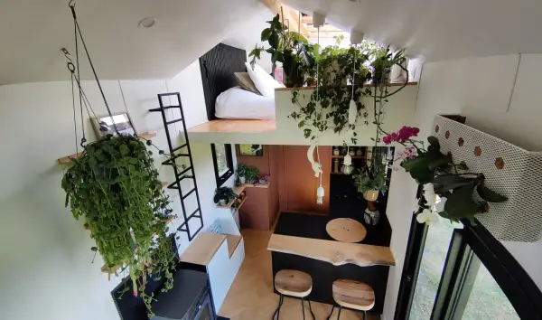 Vue aérienne du **tiny house interieur** verdoyant. Lit en mezzanine, cuisine avec bar, et cascade de plantes suspendues.