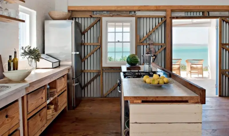 Cuisine campagne chic avec réfrigérateur, plan de travail en bois brut, murs en tôle ondulée et vue sur la mer.